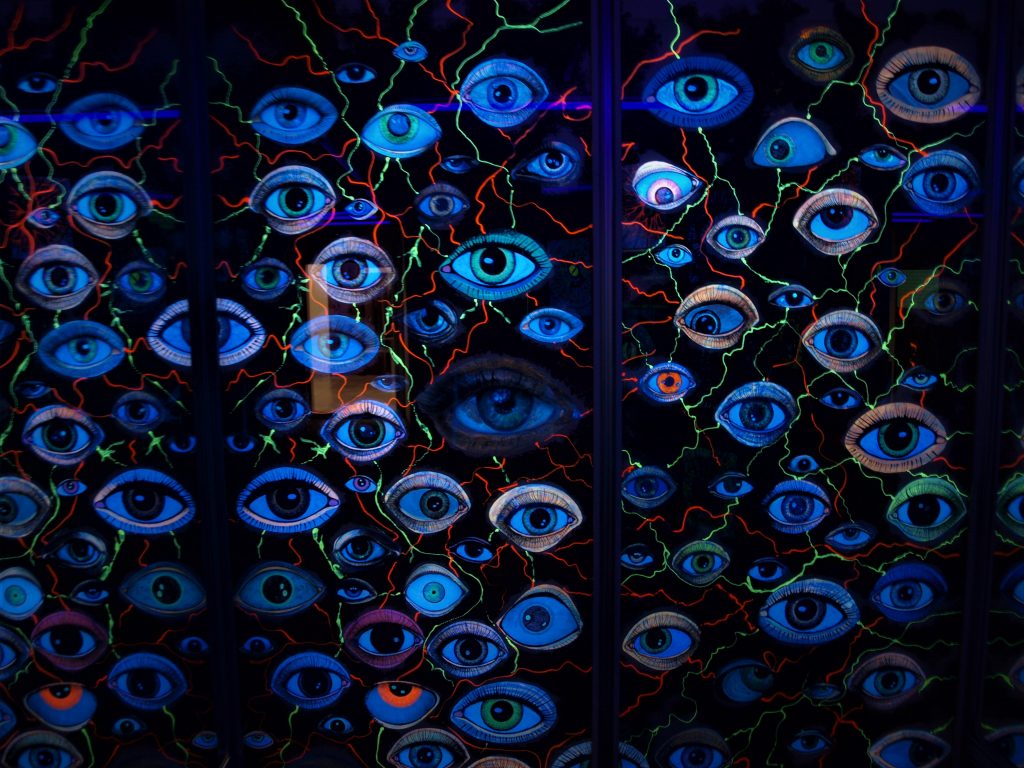 Collage con decenas de ojos en luz de neón y colores fluor contrastan con el fondo oscuro